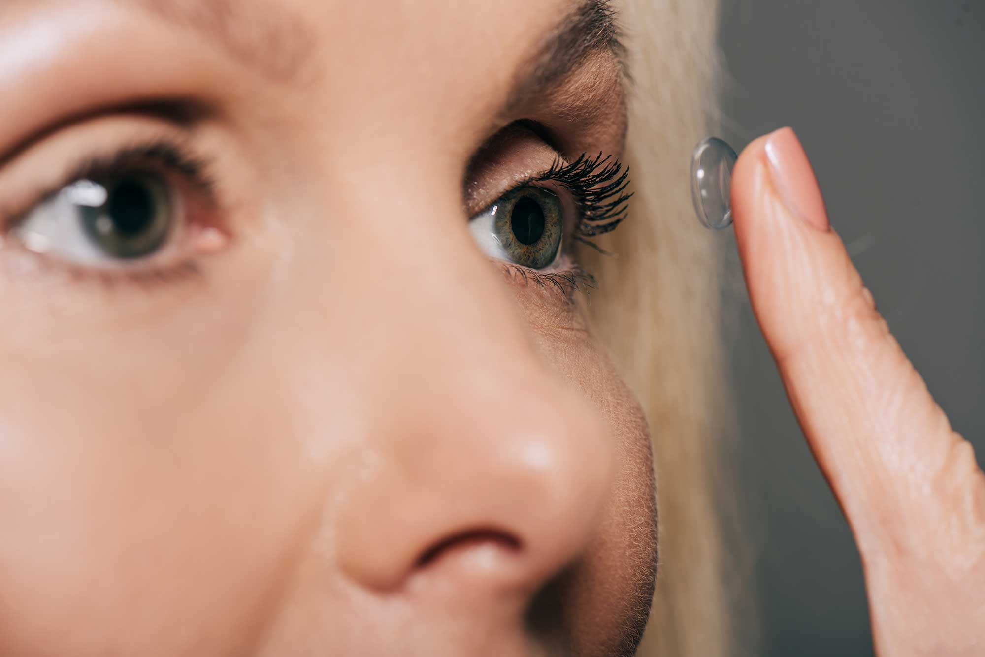 A woman holds a contact lens up to the camera, showcasing its clarity and size against a neutral background.