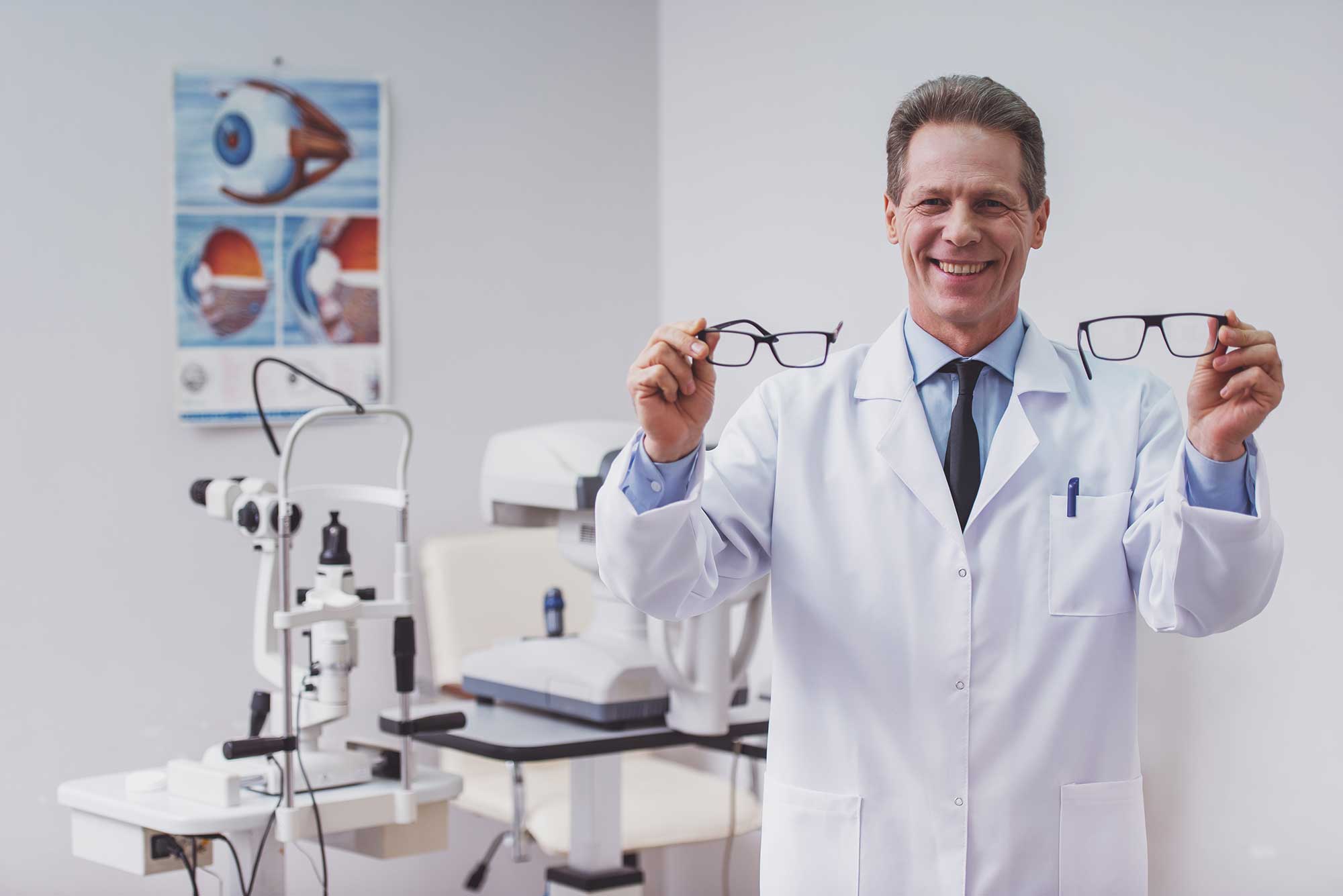 Smiling optometrist in a white coat holds two pairs of glasses in an exam room with eye test equipment and an eye anatomy poster. Professional and welcoming.